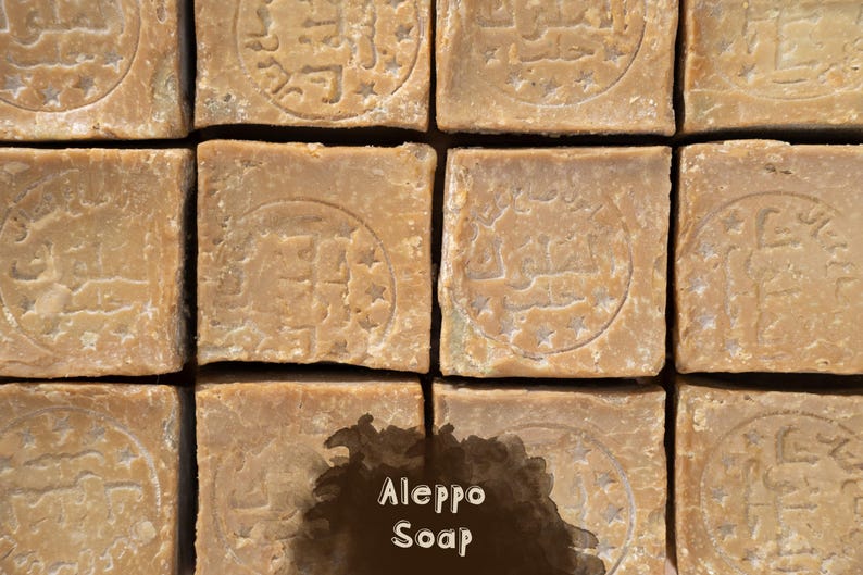 May include: A grid of square, light brown Aleppo soap bars, each featuring a circular stamped emblem with Arabic calligraphy and small stars. A dark brown, irregular watercolor-style patch is overlaid at the bottom, displaying the text Aleppo Soap.