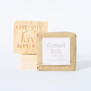 May include: Two square bars of beige soap. One bar is unwrapped, featuring an embossed camel silhouette and the text "DEVECUTLU CAMEL MILK". The second bar is wrapped in brown paper with a white label reading "Camel Milk" and ingredient text.