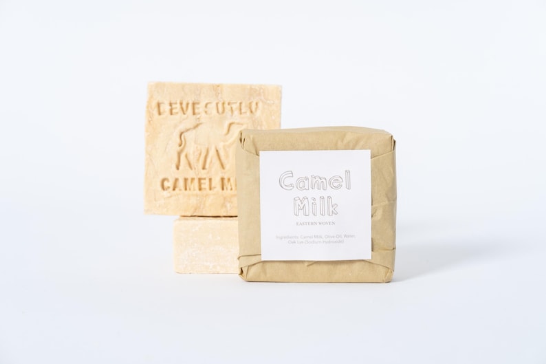 May include: Two square bars of beige soap. One bar is unwrapped, featuring an embossed camel silhouette and the text "DEVECUTLU CAMEL MILK". The second bar is wrapped in brown paper with a white label reading "Camel Milk" and ingredient text.