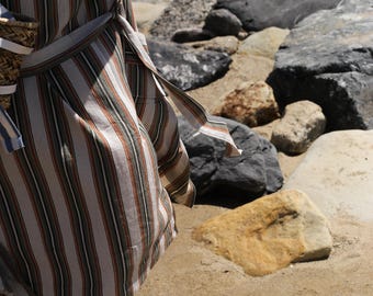Op de afbeelding: Een vrouw draagt een gestreept, lang mouwig shirt met een riem. Het shirt is lichtbruin en wit met dunne rode en groene strepen. De vrouw staat op een zandstrand met rotsen op de achtergrond.