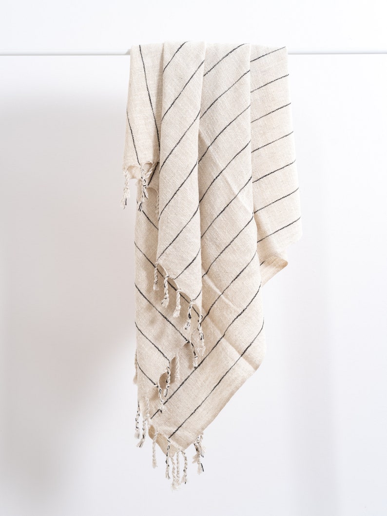 May include: A cream-colored throw blanket with black stripes and fringe. The blanket is folded and hanging from a white rod.