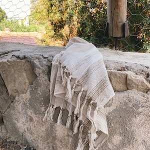 May include: A beige and brown striped throw blanket with fringe tassels. The blanket is folded and draped over a stone wall.