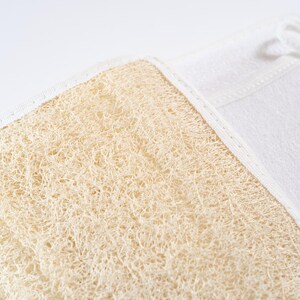 Op de afbeelding: Een close-up van een natuurlijke loofah spons bevestigd aan een witte stoffen rug. De loofah is lichtbruin van kleur en heeft een ruwe textuur.