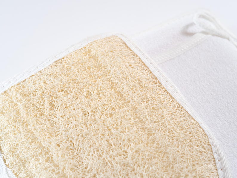 Op de afbeelding: Een close-up van een natuurlijke loofah spons bevestigd aan een witte stoffen rug. De loofah is lichtbruin van kleur en heeft een ruwe textuur.