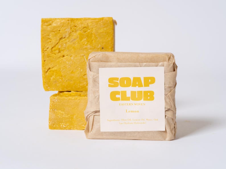 May include: Two square bars of yellow soap with a textured surface, one unwrapped and one wrapped in brown paper with a white label. The label features the yellow text SOAP CLUB, EASTERN WOVEN, Lemon, and a list of ingredients in small print.