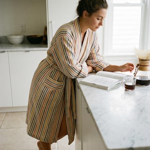 May include: A striped robe in shades of beige, red, yellow, and blue. The robe has a matching belt and pockets. A person is leaning on a countertop, reading a book, with a cup of coffee and a pour-over coffee maker nearby.