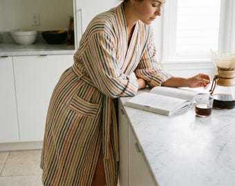 May include: A striped robe in shades of beige, red, yellow, and blue. The robe has a matching belt and pockets. A person is leaning on a countertop, reading a book, with a cup of coffee and a pour-over coffee maker nearby.