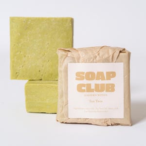 May include: Two square bars of light green soap stacked vertically, and one rectangular bar wrapped in brown paper with a white label that reads SOAP CLUB EASTERN WOVEN Tea Tree, listing ingredients including Olive Oil, Tea Tree Oil, Water, Lye.