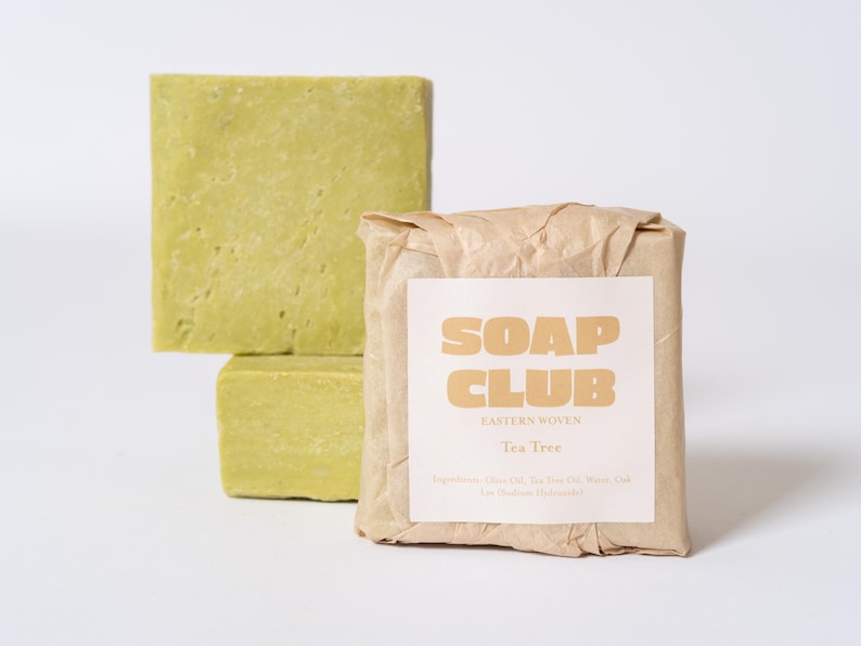 May include: Two square bars of light green soap stacked vertically, and one rectangular bar wrapped in brown paper with a white label that reads SOAP CLUB EASTERN WOVEN Tea Tree, listing ingredients including Olive Oil, Tea Tree Oil, Water, Lye.