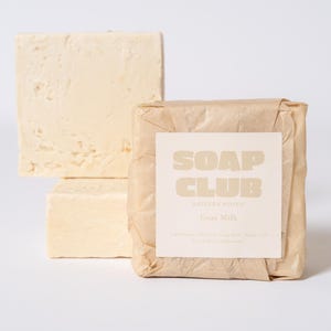 May include: Three rectangular bars of off-white goat milk soap. One bar is wrapped in crinkled brown paper with a white label that reads SOAP CLUB, EASTERN ROVEN, Goat Milk, and lists ingredients in small text. Two unwrapped bars are stacked behind.