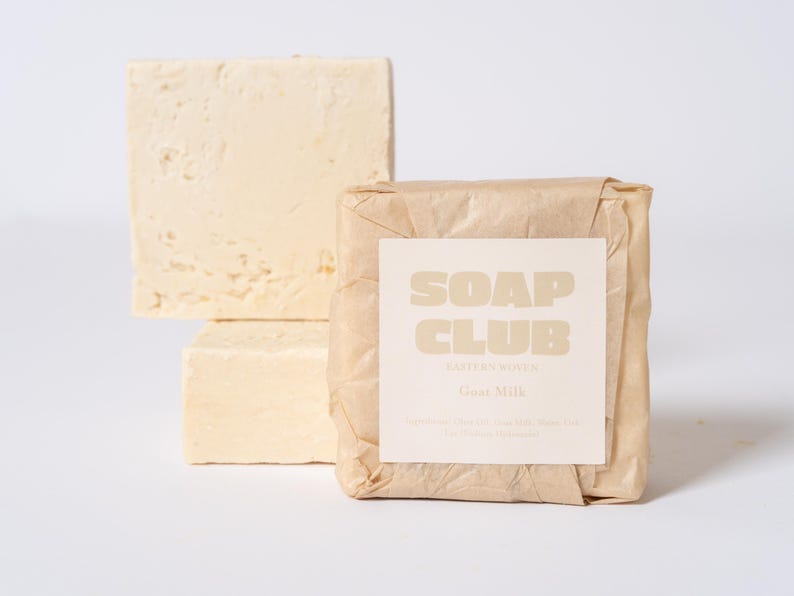 May include: Three rectangular bars of off-white goat milk soap. One bar is wrapped in crinkled brown paper with a white label that reads SOAP CLUB, EASTERN ROVEN, Goat Milk, and lists ingredients in small text. Two unwrapped bars are stacked behind.
