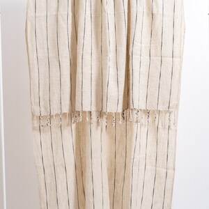 May include: A cream-colored throw blanket with black stripes and fringe. The blanket is hanging on a white rack.