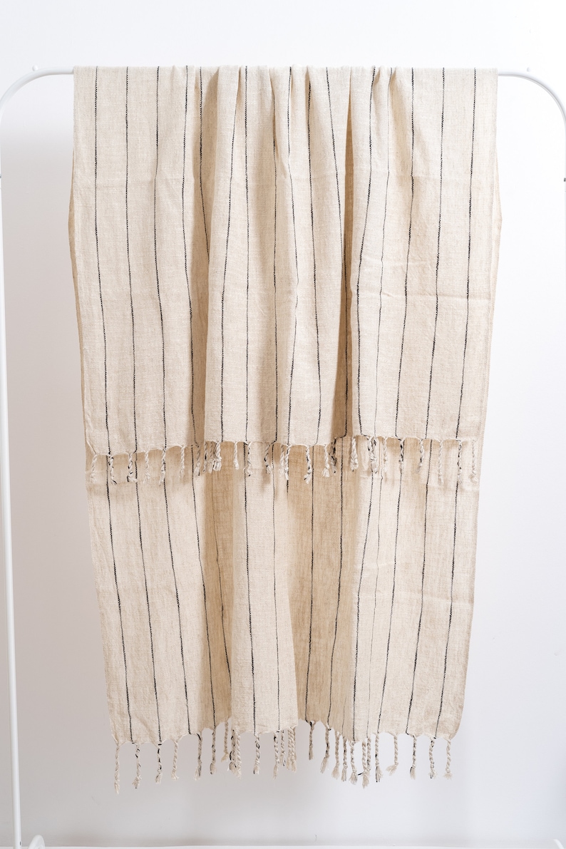 May include: A cream-colored throw blanket with black stripes and fringe. The blanket is hanging on a white rack.
