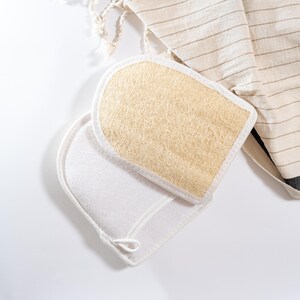Op de afbeelding: Twee scrubhandschoenen voor in bad, een witte en een beige, met een natuurlijke loofah-textuur. De handschoenen liggen op een wit oppervlak.