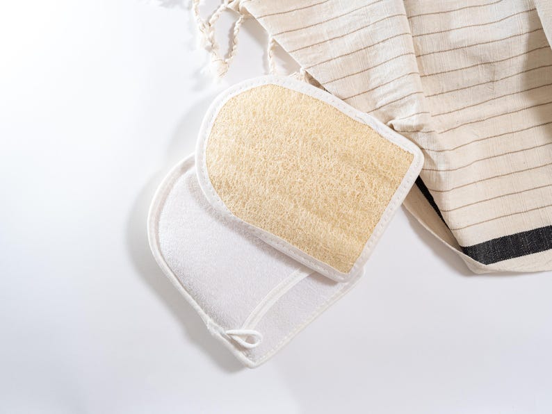 Op de afbeelding: Twee scrubhandschoenen voor in bad, een witte en een beige, met een natuurlijke loofah-textuur. De handschoenen liggen op een wit oppervlak.