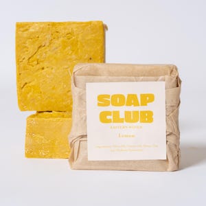 Puede incluir: Dos barras de jabón amarillas, una apilada encima de la otra, junto a una barra envuelta. La barra envuelta tiene una etiqueta que dice "SOAP CLUB" y "Lemon". Las barras de jabón son de color amarillo brillante.