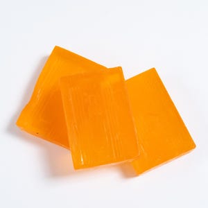 May include: Three rectangular bars of translucent orange soap arranged in a slightly overlapping cluster. Each bar features a smooth, glossy surface with subtle vertical lines embossed into the material, creating a uniform appearance across all pieces.