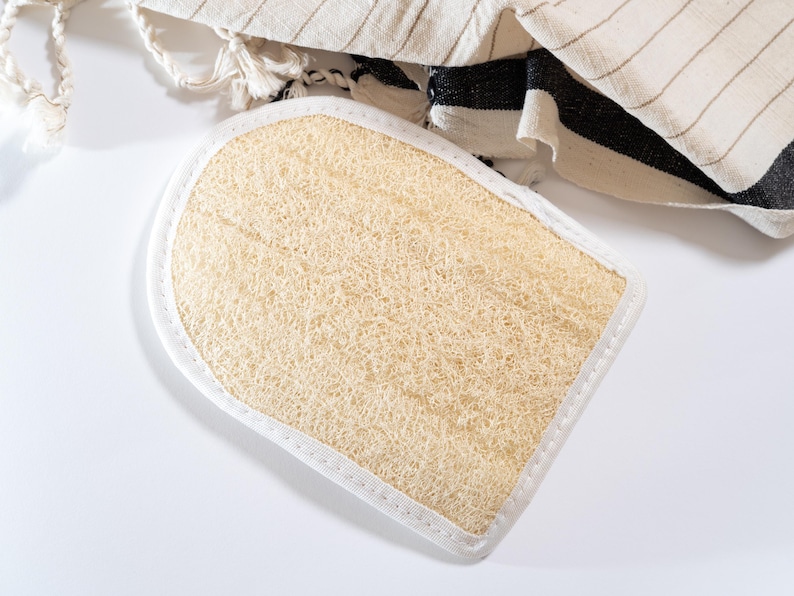 Op de afbeelding: Een beige loofah badspons met een witte gestikte rand. De spons is ovaalvormig en heeft een textuur.