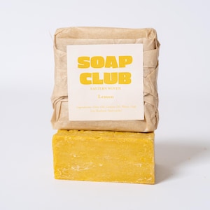May include: A rectangular bar of yellow soap positioned below a second bar of soap wrapped in light brown paper. The paper wrapper features a white label with the yellow text SOAP CLUB, EASTERN WOVEN, and Lemon, along with a list of ingredients.