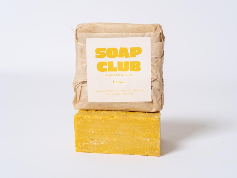 May include: A rectangular bar of yellow soap positioned below a second bar of soap wrapped in light brown paper. The paper wrapper features a white label with the yellow text SOAP CLUB, EASTERN WOVEN, and Lemon, along with a list of ingredients.