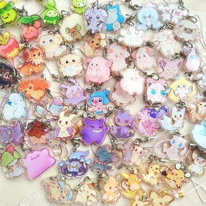 May include: A collection of numerous small, clear acrylic charms featuring colourful cartoon creature illustrations. Each charm is attached to a thin, flexible cord loop, showcasing a variety of pastel and vibrant characters in diverse poses.
