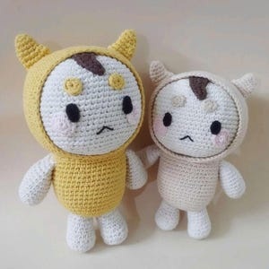 Puede incluir: Dos muñecos hechos a mano con ganchillo. Un muñeco lleva un conjunto con capucha amarillo y el otro un conjunto con capucha color crema. Ambos muñecos tienen caras blancas con ojos negros, una mancha marrón y mejillas rosas. Los muñecos tienen brazos y piernas pequeños.