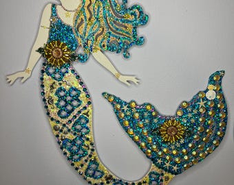 May include: A mermaid silhouette made of wood and decorated with iridescent blue and gold sequins, beads, and other embellishments. The mermaid's hair is long and flowing, and her tail is curled around her body.