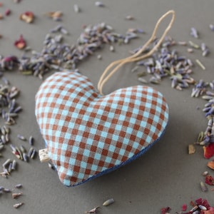 heart shaped organic lavender sachet, 3 inch tall, made out of upcycled cotton with light blue and brown gingham. Sachet has a twine loop for hanging, and is displayed with loose lavender and rose petals around it.