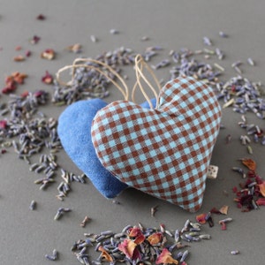 2 heart shaped organic lavender sachets, 3 inch tall, made out of upcycled cotton with light blue and brown gingham, and light denim back. Sachets have a twine loop for hanging, and are displayed with loose lavender and rose petals around it.