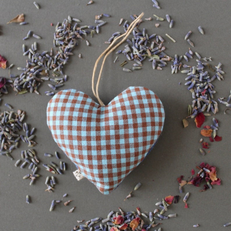 heart shaped organic lavender sachet, 3 inch tall, made out of upcycled cotton with light blue and brown gingham. Sachet has a twine loop for hanging, and is displayed with loose lavender and rose petals around it. close up view.