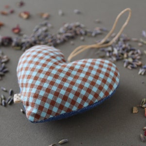 heart shaped organic lavender sachet, 3 inch tall, made out of upcycled cotton with light blue and brown gingham. Sachet has a twine loop for hanging, and is displayed with loose lavender and rose petals around it. Side view.