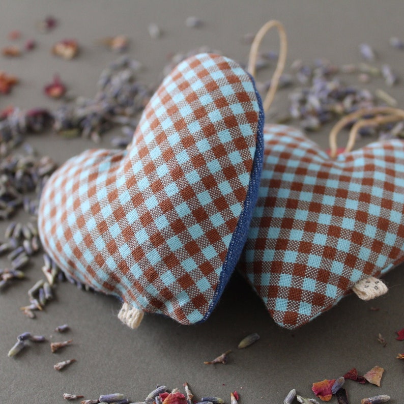 two heart shaped organic lavender sachets, 3 inch tall, made out of upcycled cotton with light blue and brown gingham. Sachets have a twine loop for hanging, and are displayed with loose lavender and rose petals around it.