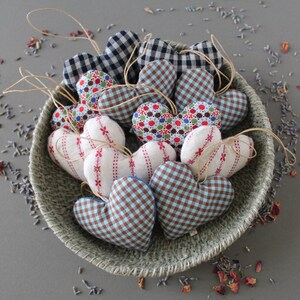 heart shaped organic lavender sachet, 3 inch tall, made out of cotton with light blue and brown gingham. Sachet has a twine loop for hanging, and is displayed in a basket with other sachets of different colors, and loose lavender and rose petals.