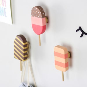 Peut inclure: Crochets muraux en bois en forme de glace, de couleurs marron, rose et jaune, avec un bâton en bois clair. Une décoration d'œil noir et une pancarte avec l'inscription "SUN SHINE" sont également visibles.