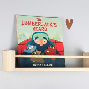 May include: A wooden floating shelf holds a children's book titled "The Lumberjack's Beard" with illustrations of a lumberjack and forest animals. The book is on a light gray wall with brown heart-shaped decorations.