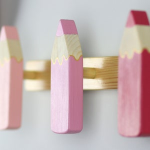 May include: Three wooden pencil-shaped hooks in pink and red colors with gold accents. The hooks are mounted on a wooden board.
