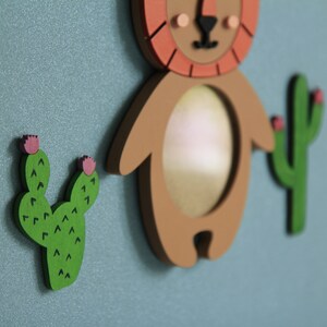 May include: A brown wooden lion with a red mane and a round mirror in the center. The lion is surrounded by two green cactus plants with pink flowers.