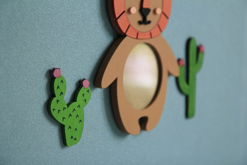 May include: A brown wooden lion with a red mane and a round mirror in the center. The lion is surrounded by two green cactus plants with pink flowers.