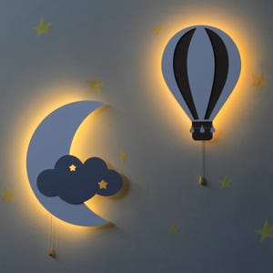 Könnte beinhalten: Zwei weiße Wandleuchten mit einer hellblauen Wolke und einem Heißluftballon-Design. Die Leuchten sind mit warmweißem Licht beleuchtet.