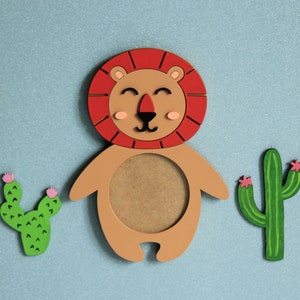 May include: A wooden lion picture frame with a brown background and a red mane. The frame is surrounded by two green cactus plants with pink flowers.