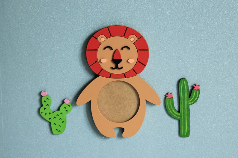 May include: A wooden lion picture frame with a brown background and a red mane. The frame is surrounded by two green cactus plants with pink flowers.