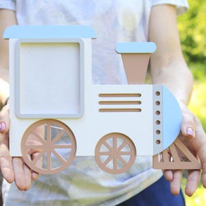 Op de afbeelding: Een witte en blauwe houten trein met een fotolijst aan de voorkant. De trein heeft bruine wielen en een bruine schoorsteen. De trein wordt in de handen van een persoon gehouden.