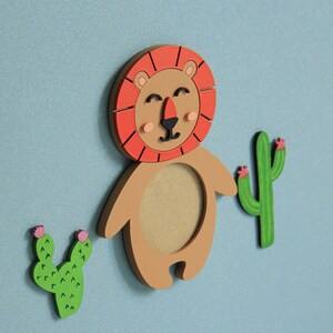 May include: A wooden lion figurine with a red mane and a brown body. The lion has a round cutout in its chest. Two green cactus figurines with pink flowers are on either side of the lion.