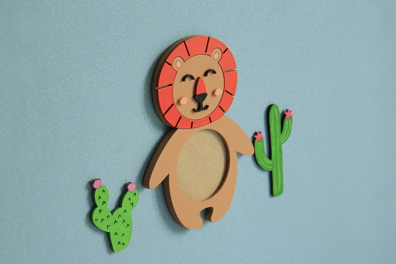 May include: A wooden lion figurine with a red mane and a brown body. The lion has a round cutout in its chest. Two green cactus figurines with pink flowers are on either side of the lion.