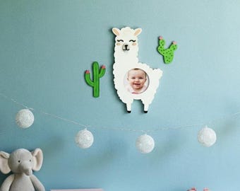 May include: A white llama-shaped picture frame with a baby's photo, flanked by green cactus decorations on a light blue wall. A string of decorative white globe lights hangs below. A stuffed elephant, books, and pink shoes are on a white shelf.