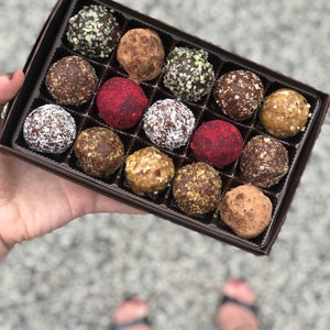 May include: A box of assorted energy balls, with flavors including chocolate, coconut, and fruit. The balls are arranged in a grid pattern in a brown box.