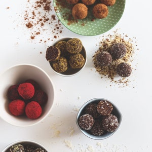 May include: Five white bowls filled with various flavors of homemade energy balls. The bowls are on a white surface. The energy balls are covered in different toppings, including coconut flakes, cocoa powder, and nuts.