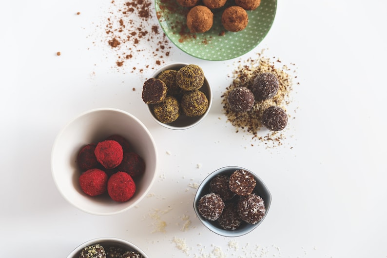 May include: Five white bowls filled with various flavors of homemade energy balls. The bowls are on a white surface. The energy balls are covered in different toppings, including coconut flakes, cocoa powder, and nuts.
