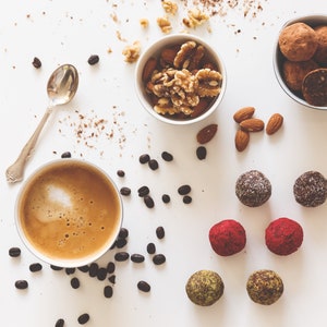 May include: A white surface with a cup of coffee, a bowl of walnuts and almonds, a bowl of chocolate truffles, and a variety of colorful energy balls.