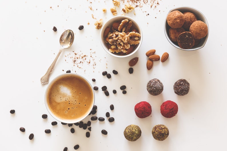 May include: A white surface with a cup of coffee, a bowl of walnuts and almonds, a bowl of chocolate truffles, and a variety of colorful energy balls.